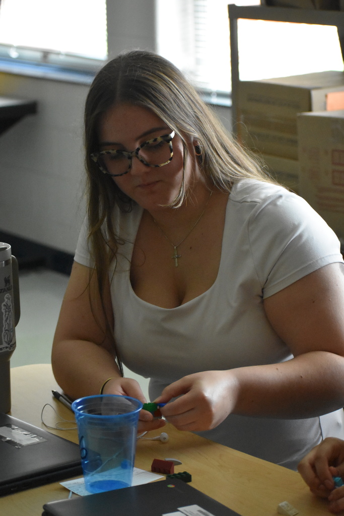 A student with long light brown hair and tortoiseshell glasses, wearing a white shirt and a cross necklace, sits at a desk working with green and blue Lego pieces. A blue plastic cup, Chromebook, and water bottle are nearby.