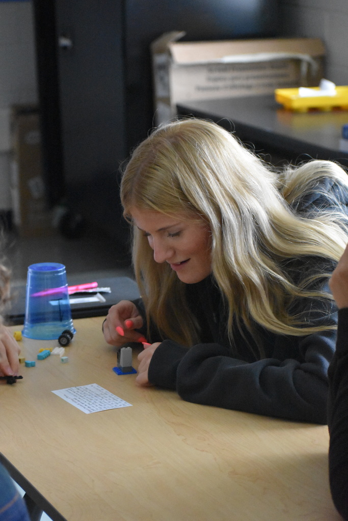A student with long blonde hair, wearing a black hoodie, leans over a desk and smiles while assembling Lego pieces. A blue plastic cup, small Lego parts, and a handwritten note are on the table.