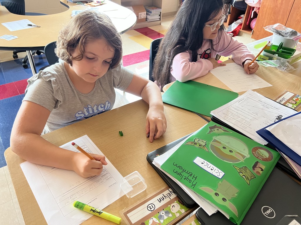 Two students sit side by side at a desk, each writing on a science worksheet. One student has short hair and a gray Stitch shirt, the other has long dark hair and glasses. A green folder with Baby Yoda stickers and a Chromebook are on the desk.