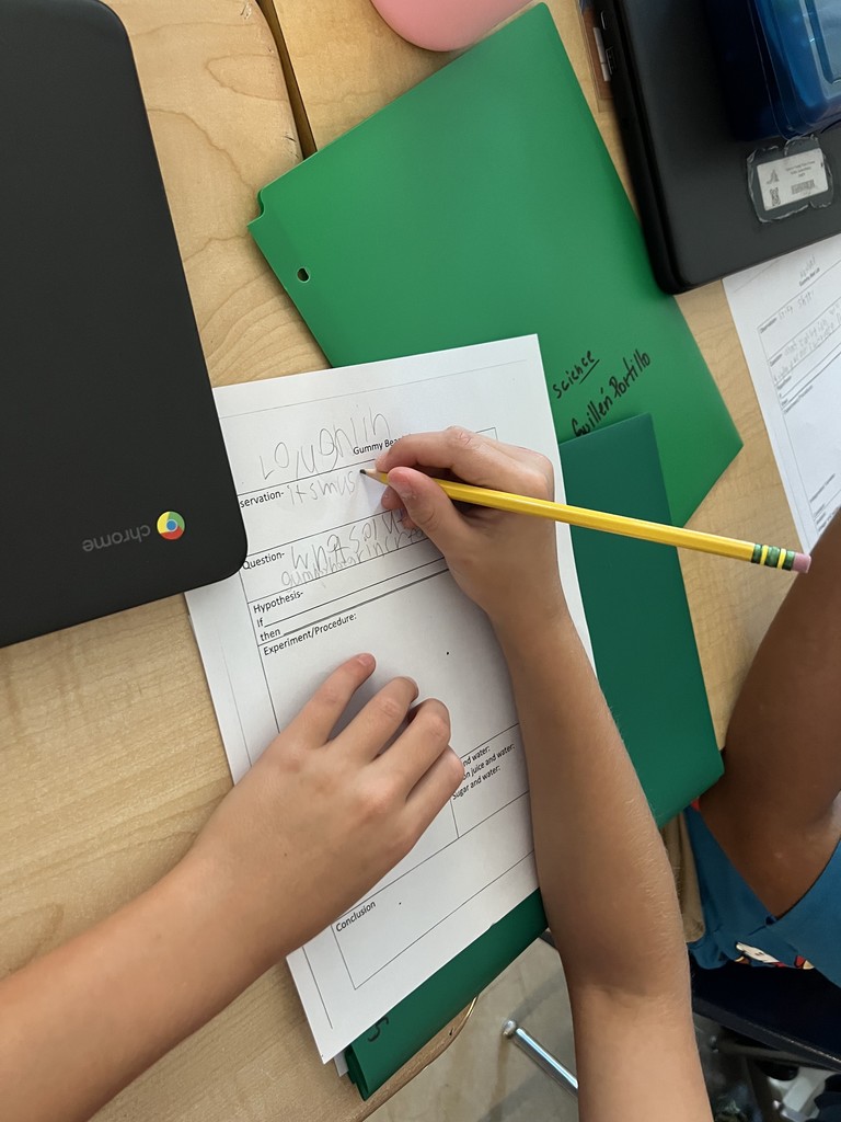 A student writes on a science worksheet titled “Gummy Bear” with a pencil, while another hand rests on the desk. A Chromebook, green folder, and pink mouse are nearby.