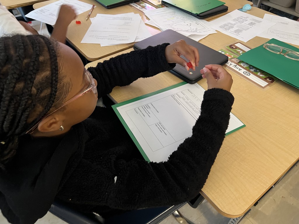 A student with braided hair and glasses sits at a desk, holding a red gummy bear above a science worksheet inside a green folder. Papers, a Chromebook, and other supplies are spread across the table.