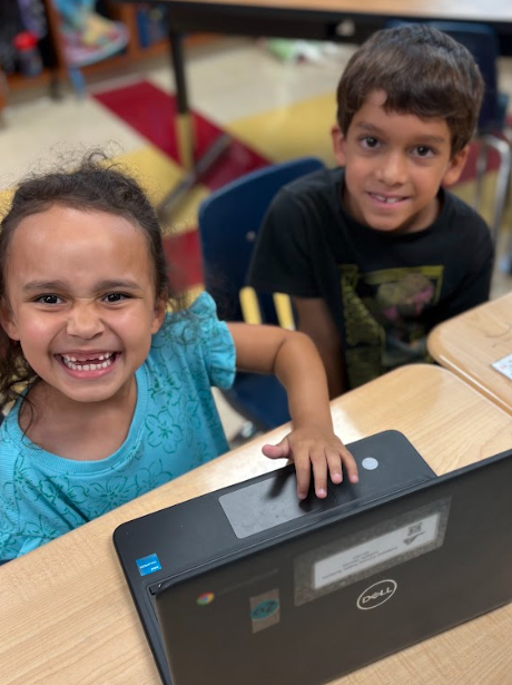 Two young students sitting at a desk smiling at the camera, with a laptop open in front of them.