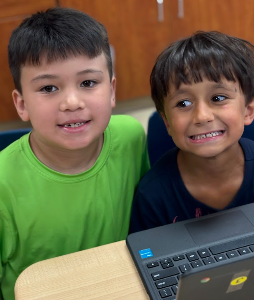 wo boys sitting next to each other at a desk, smiling with a laptop in front of them.