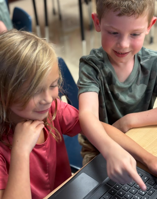 A boy pointing at a laptop screen while a girl next to him watches and smiles.