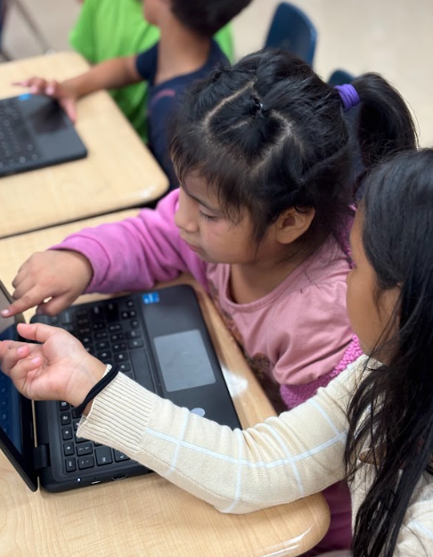 Two girls working together on a laptop, one pointing at the screen while the other observes closely.