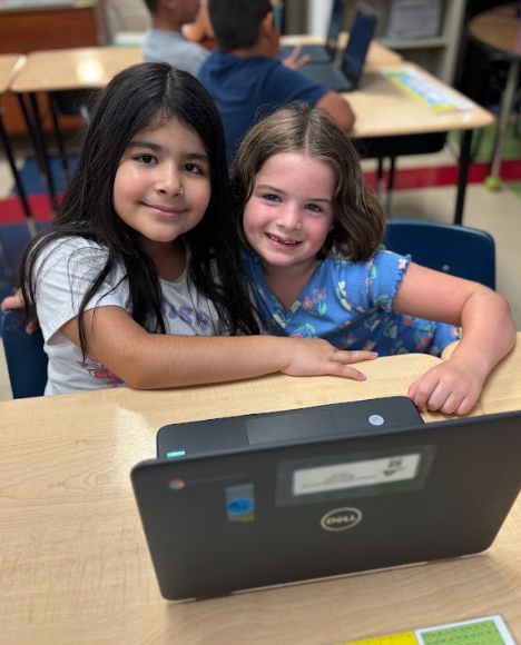 Two girls sitting close together at a desk, smiling and posing in front of a laptop.