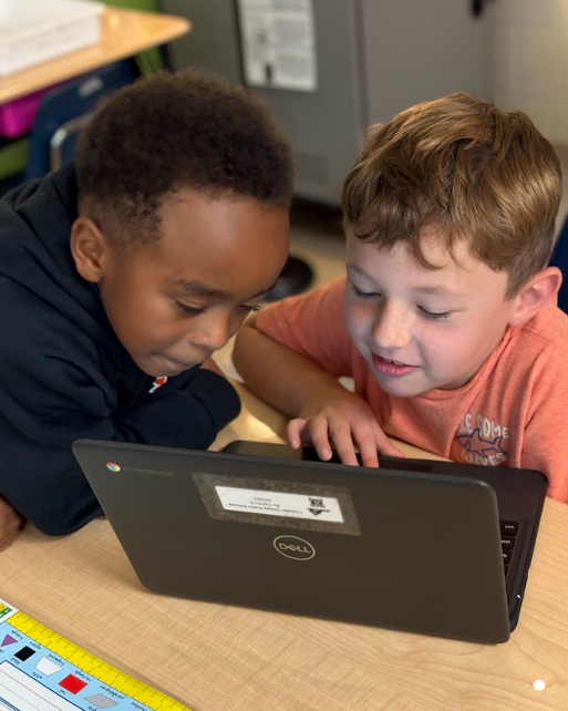 Two boys leaning over a laptop at a desk, concentrating as they look at the screen together.