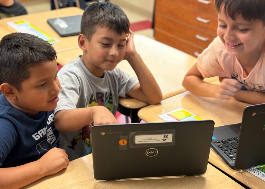 Three boys gathered around a laptop at a desk, one pointing at the screen while the others watch and smile.
