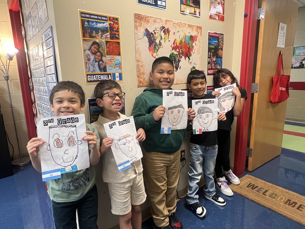 Group of five students holding portraits – Five smiling elementary students stand in a hallway holding self-portraits labeled “3rd Grade.” Behind them are posters and a colorful world map.