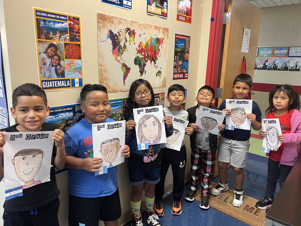 Line of seven students with portraits – Seven elementary students stand side by side in a hallway, each holding a “2nd Grade” self-portrait. A large map of the world and cultural posters are displayed on the wall behind them.