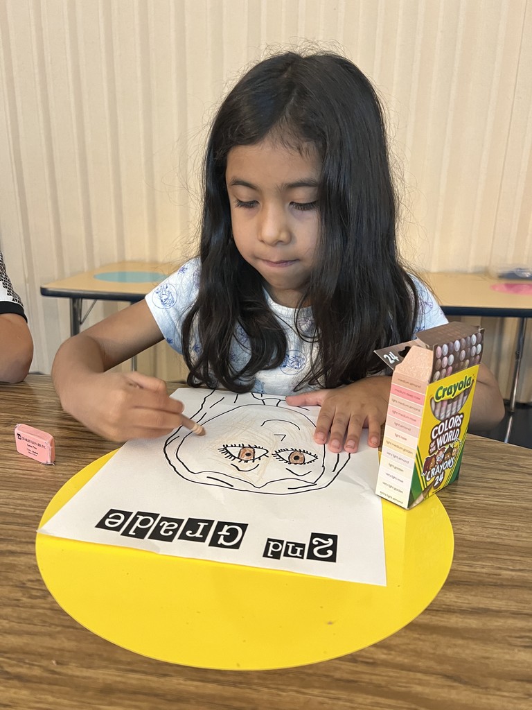 A girl with long dark hair carefully colors her “2nd Grade” self-portrait at a classroom desk. A box of Crayola “Colors of the World” crayons is on the table.