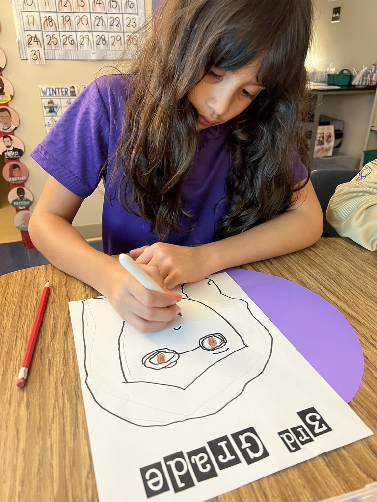 A student with long dark hair in a purple shirt leans over her paper while drawing a “3rd Grade” self-portrait with markers and pencils at a classroom desk.
