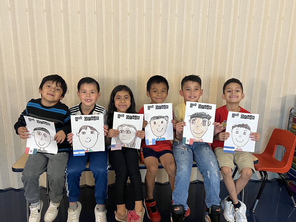Seated group with portraits – Six students sit in a row on a bench, smiling and holding “2nd Grade” self-portraits. Each portrait is different, and small flags are attached to the papers.