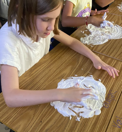 A student in a white shirt concentrates while using her hand to spread shaving cream in a circle on her desk. Another child is partially visible working at the next desk.
