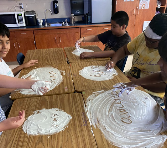 Four boys sit at a cluster of desks, each with shaving cream spread out. They write words in the foam, including one student who has written “change” in large letters.