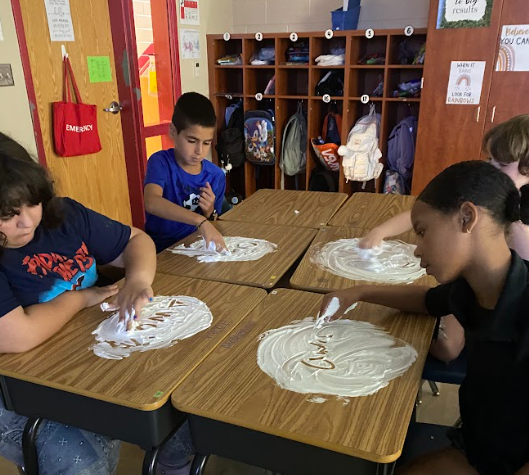 Four elementary students sit at desks spreading shaving cream across the surface and writing letters with their fingers. Behind them are cubbies filled with backpacks and supplies.