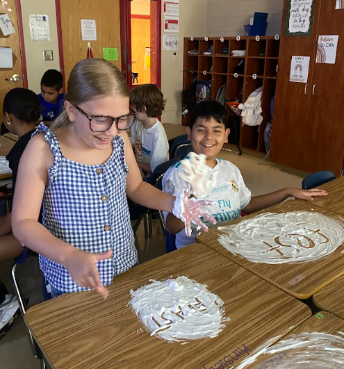Two smiling students sit side by side. The girl in a blue checkered dress and glasses has shaving cream on her hands, while the boy next to her looks at the foam on his desk where he has written “fast.”