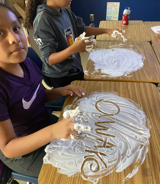 Two students work at desks covered in shaving cream. The girl in the foreground, wearing a purple Nike shirt, has written the word “awake” in the foam.