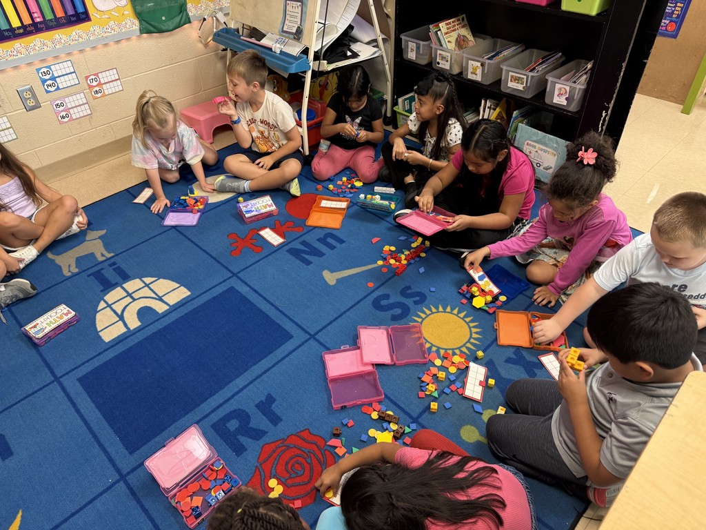 A group of students sit in a circle on a classroom rug, working with manipulatives from their math toolkits. They are using counting cubes, pattern blocks, and other colorful pieces, collaborating in small groups while exploring hands-on math activities.