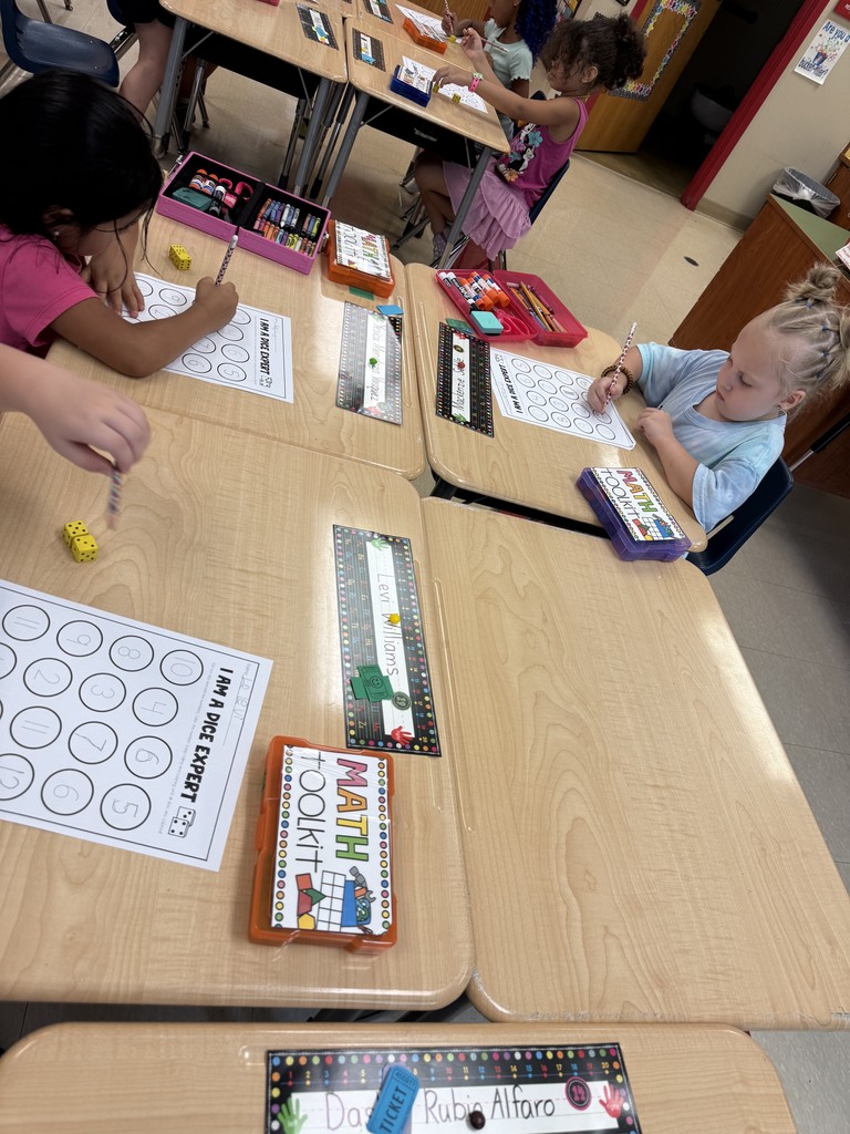 Several students sit at desks working on the same “I Am a Dice Expert” math sheet. Each student has a math toolkit box, dice, and writing supplies. The children are focused on rolling dice and writing numbers, with colorful supply boxes on their desks.