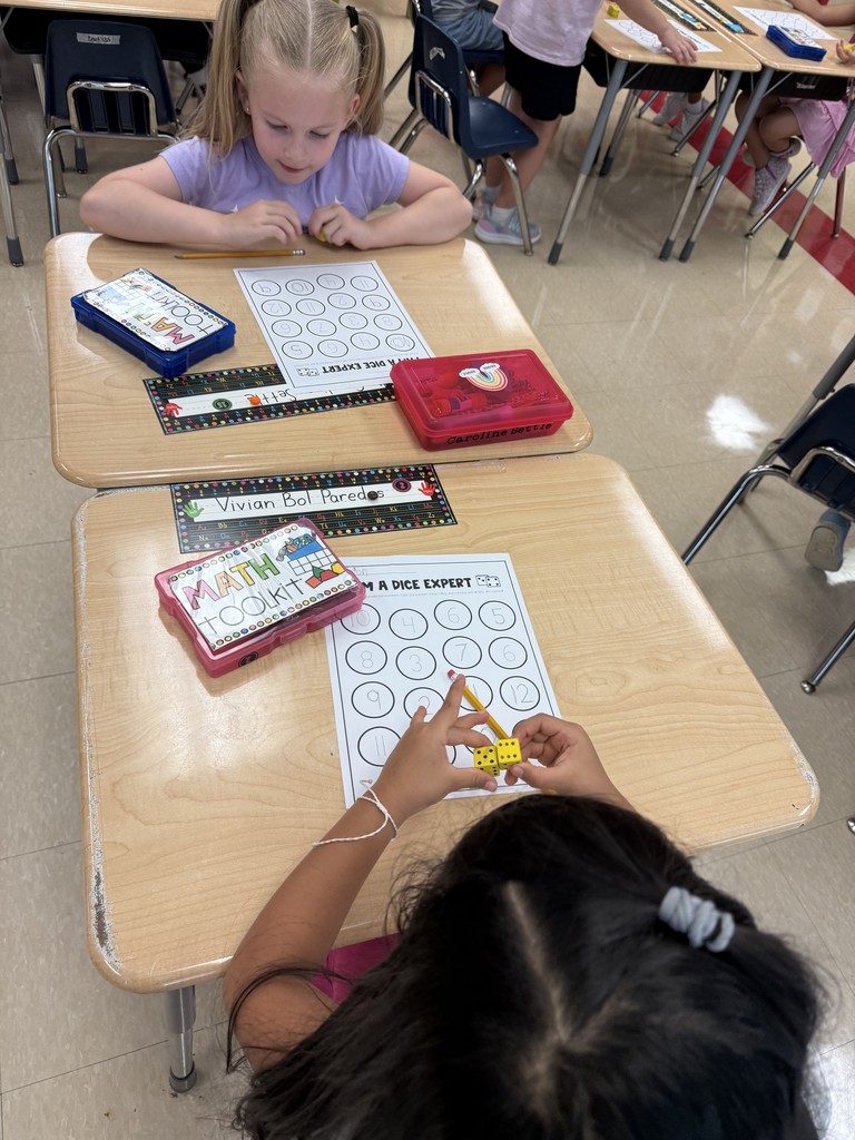 Two students work at desks on the “I Am a Dice Expert” sheet. One student rolls two yellow dice while the other looks thoughtfully at her paper. Math toolkits and pencils are on the desks, and the children are engaged in practicing number recognition and addition with dice.