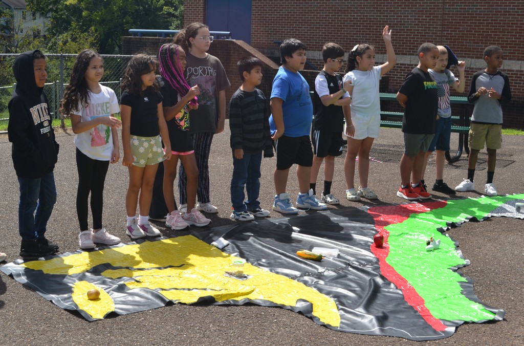 Image contains a group of students outside.  There is a large duck tape map of Virginia and students are placing products in various spots on the map.