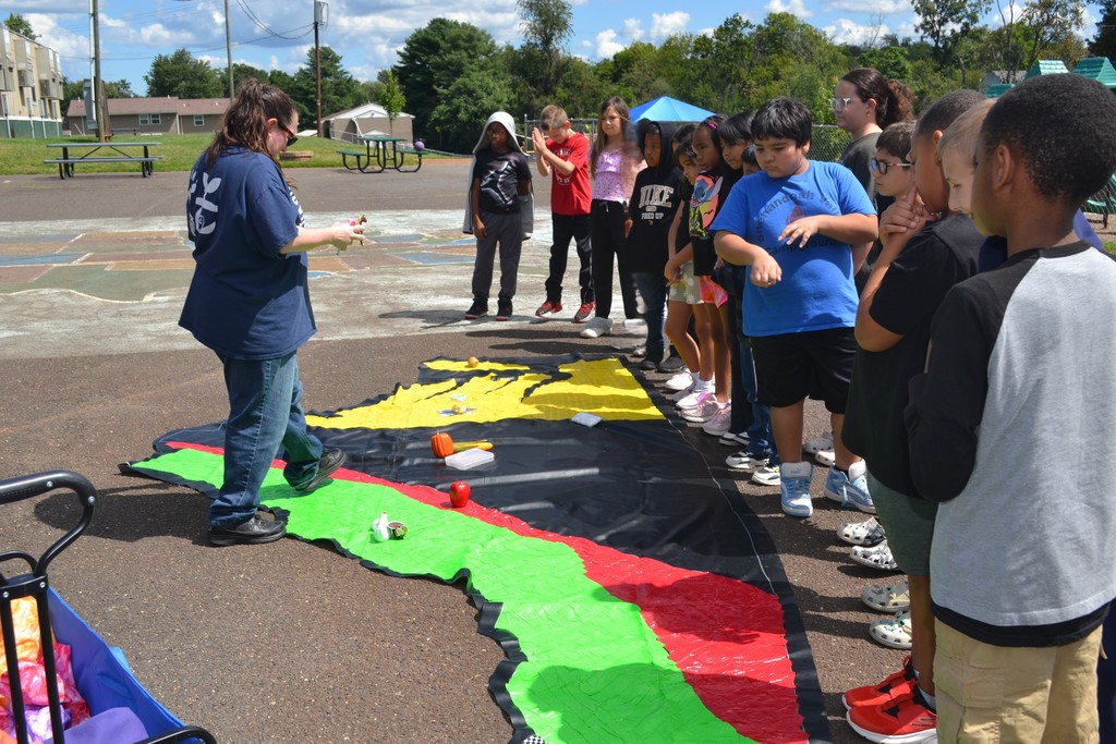 Image contains a group of students outside with the guest presenter.  There is a large duck tape map of Virginia and students are placing products in various spots on the map.