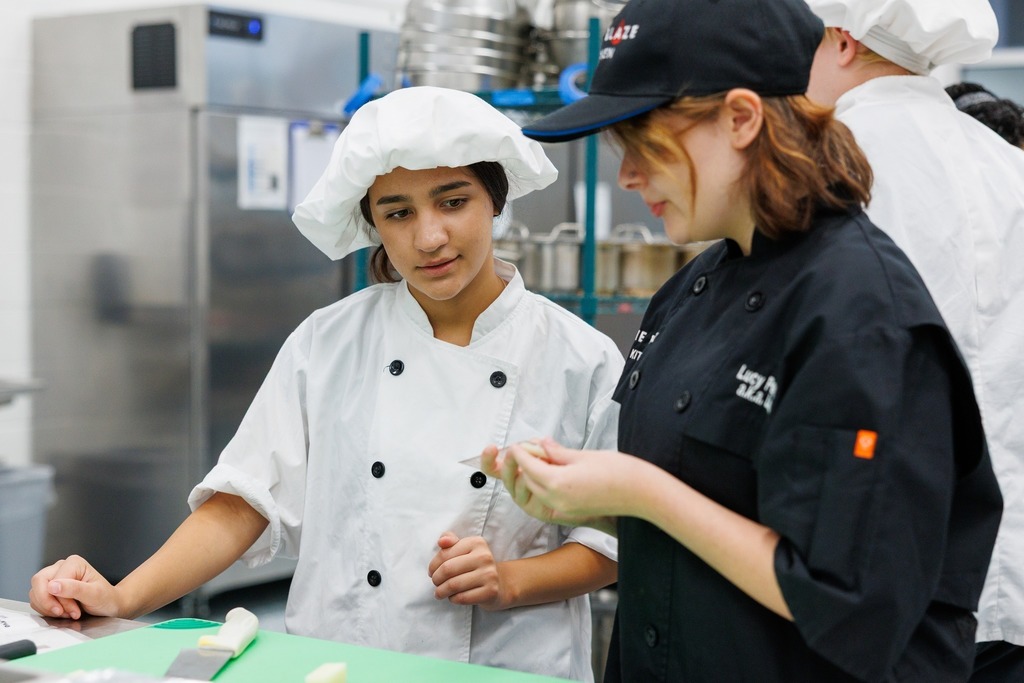 Two culinary students in a kitchen classroom. One student in a white chef coat and hat listens attentively while another student in a black chef coat and cap demonstrates a food preparation technique.