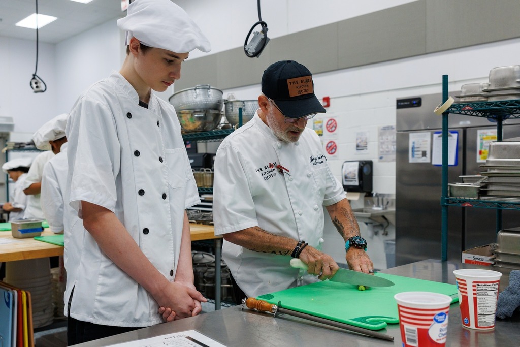 A culinary instructor in a white chef coat and black cap demonstrates knife skills on a green cutting board while a student in a white chef coat and hat observes closely in a commercial kitchen.