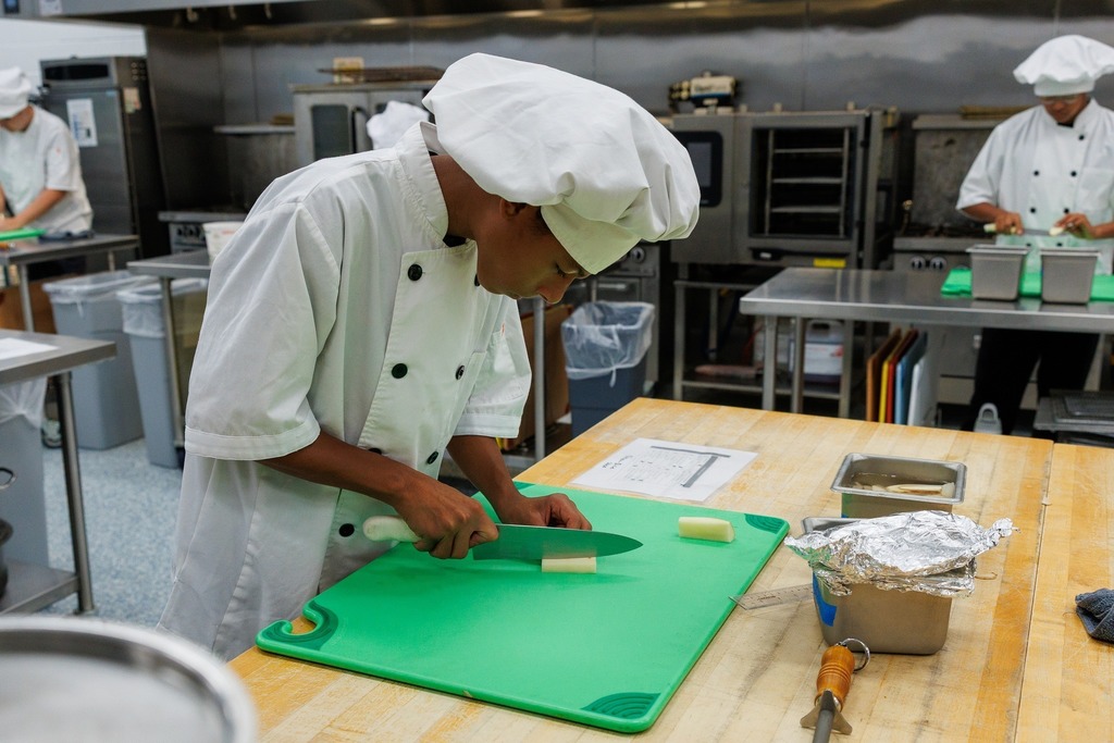 A culinary student in a white chef coat and hat slices a piece of vegetable on a green cutting board in a commercial kitchen, with ovens and other students visible in the background.