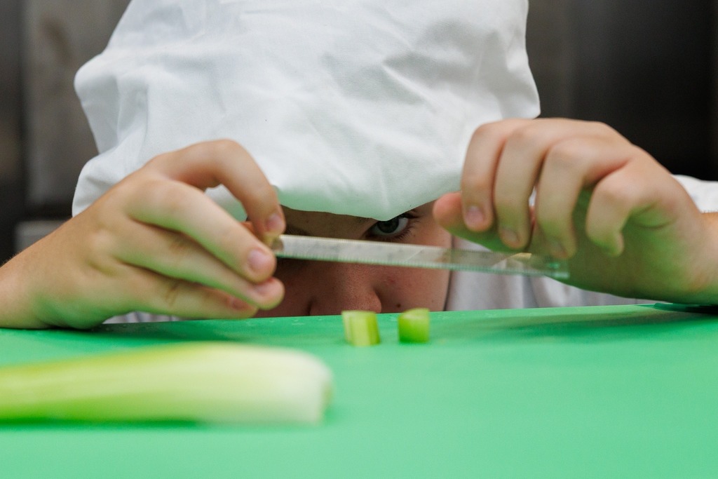 A culinary student in a white chef hat peers closely at chopped vegetable pieces on a green cutting board, measuring them with a ruler to check for uniform size.