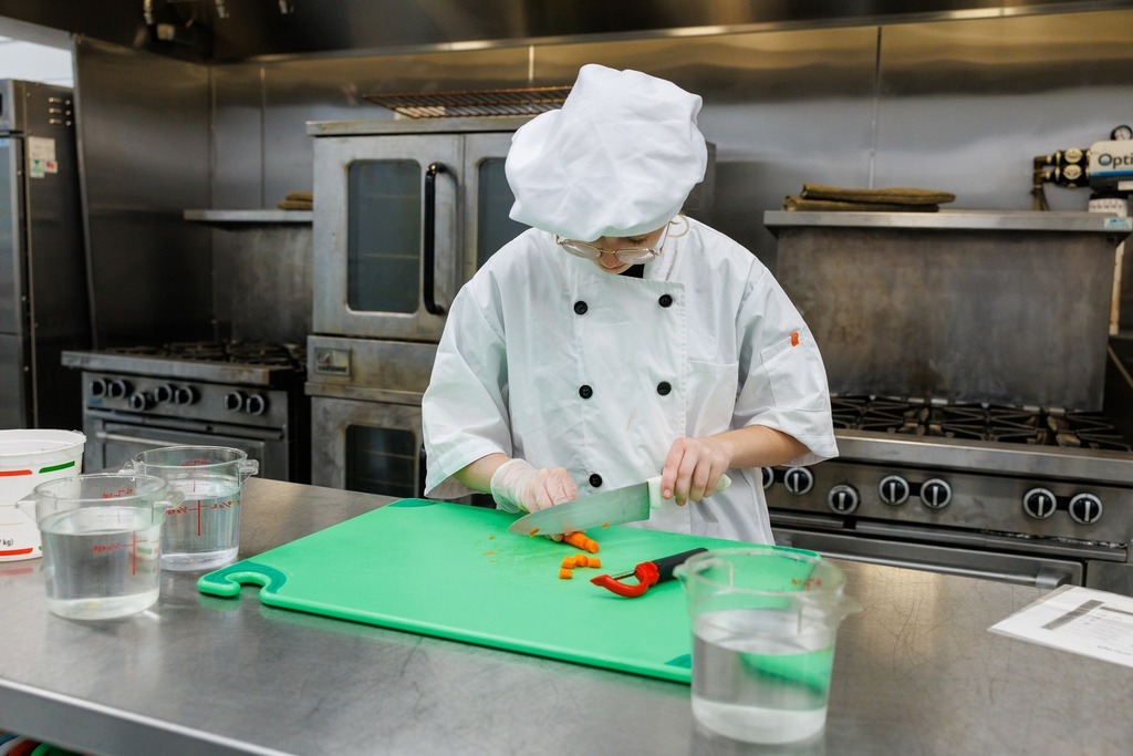 A culinary student in a white chef coat and hat chops orange vegetables on a green cutting board in a commercial kitchen, with measuring cups of water and kitchen equipment nearby.