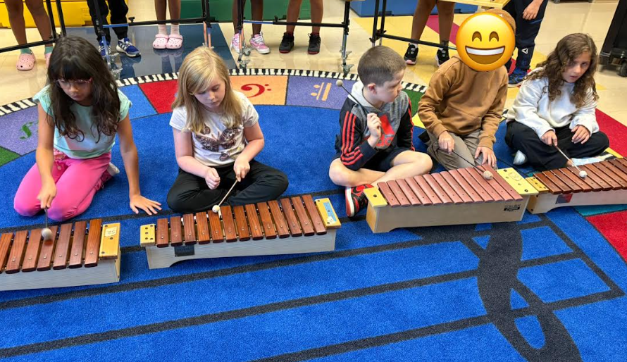 Four children sit on a blue rug decorated with music staff lines, each playing a wooden xylophone with mallets. Other students’ legs are visible in the background, standing behind them.