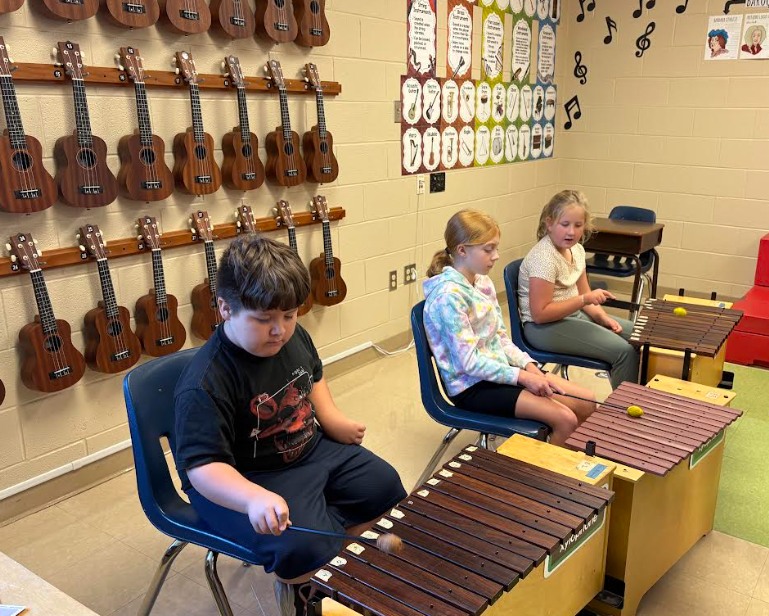 Three children sit in chairs playing xylophones. Behind them, multiple ukuleles are mounted on the classroom wall.
