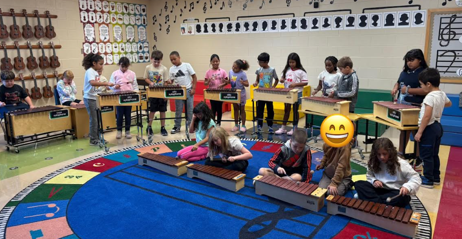 A group of children are playing xylophones in a music classroom. Some sit on the colorful rug in the front while others stand behind larger instruments. Music notes and silhouette portraits decorate the classroom wall.
