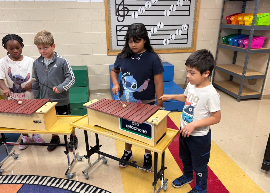 Four children stand at xylophones, playing with mallets. They are positioned on the colorful classroom floor, with music decorations and shelving in the background.