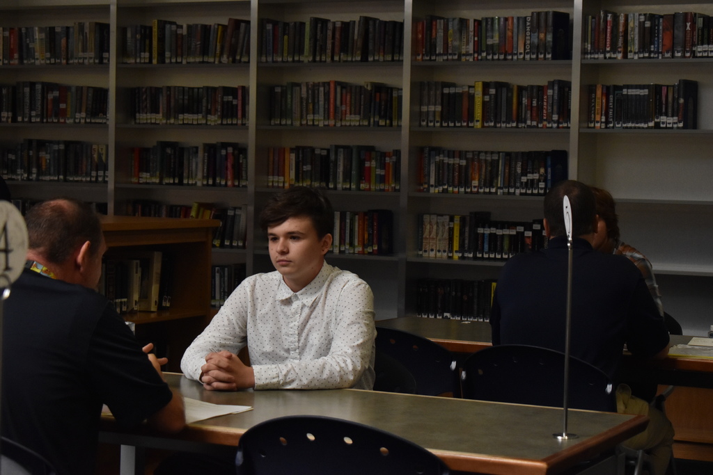 A student in a white polka dot shirt sits at a table with his hands clasped. He is looking straight ahead at the man interviewing him, engaging in direct eye contact. They are surrounded by shelves of books in a high school library.