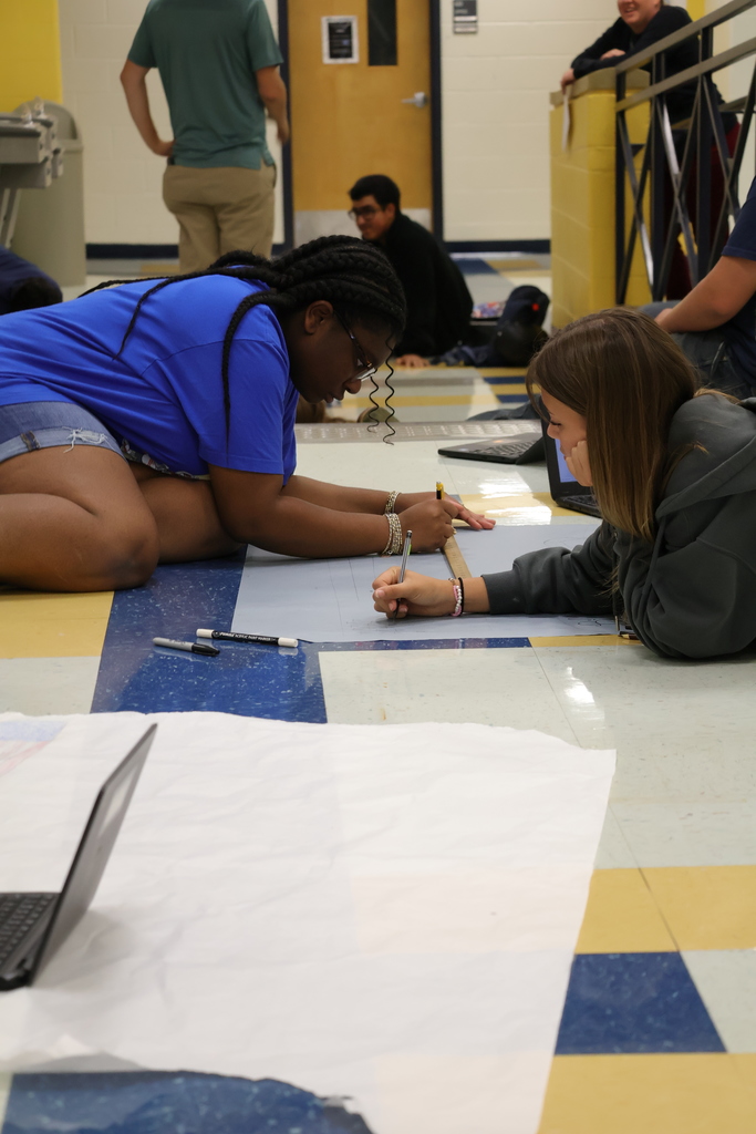 Two students work together on a large sheet of paper while lying on the floor of a school hallway. One student in a blue shirt uses a ruler while the other in a gray hoodie holds a pencil. Laptops and markers are nearby, and other students are visible in the background.