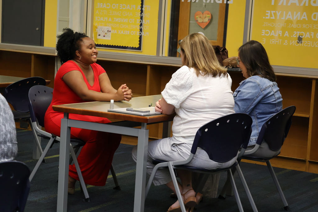 A student in a red dress smiles while sitting at a table in a high school library. Opposite her sit two interviewers in business casual attire. 