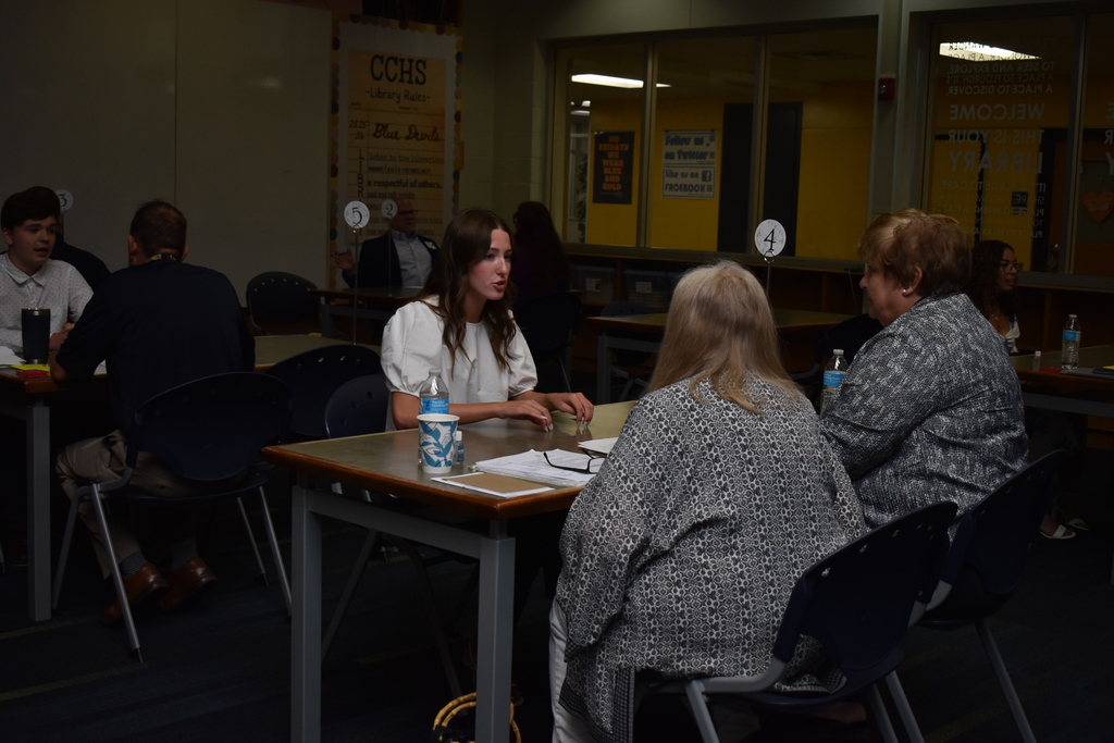 A student in a white blouse talks to two interviewers at a table in a high school library. There are waterbottles on the table with the table number four in the air. She is articulating with her hands.