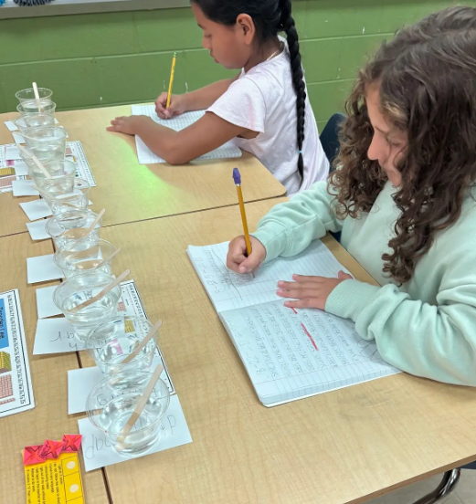 Two girls writing in notebooks at a table lined with clear cups of water labeled with different substances, part of a science investigation.
