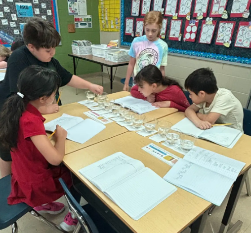 Five children gathered around a table with a line of clear cups filled with water, writing notes and testing with pH strips.