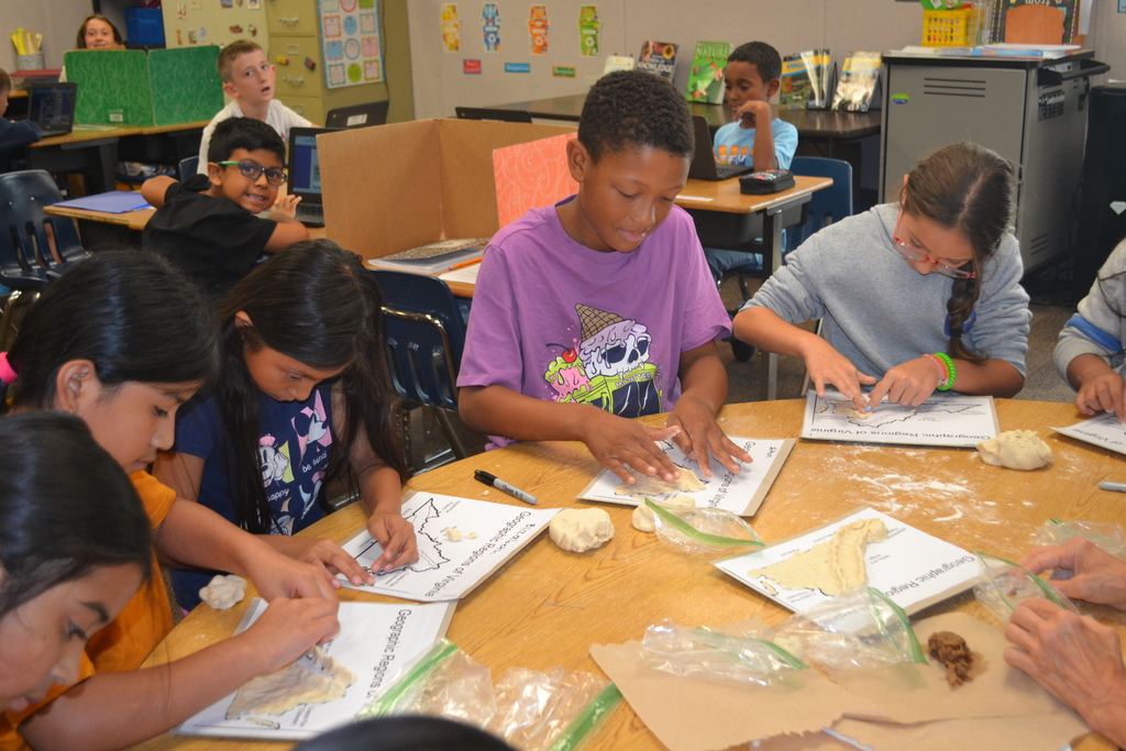 A group of students gather around a table working on a salt dough map of Virginia.  Students in the background are working on their computers.