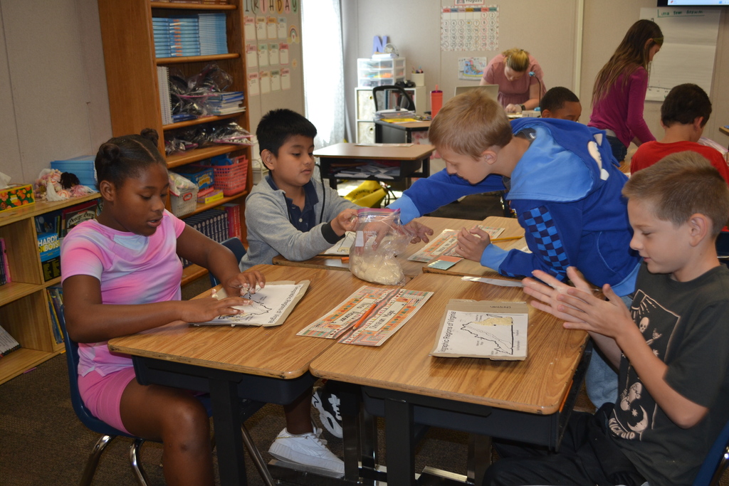 A group of 4 students are working on a salt dough map of Virginia at their desks.  Teacher in the far background.