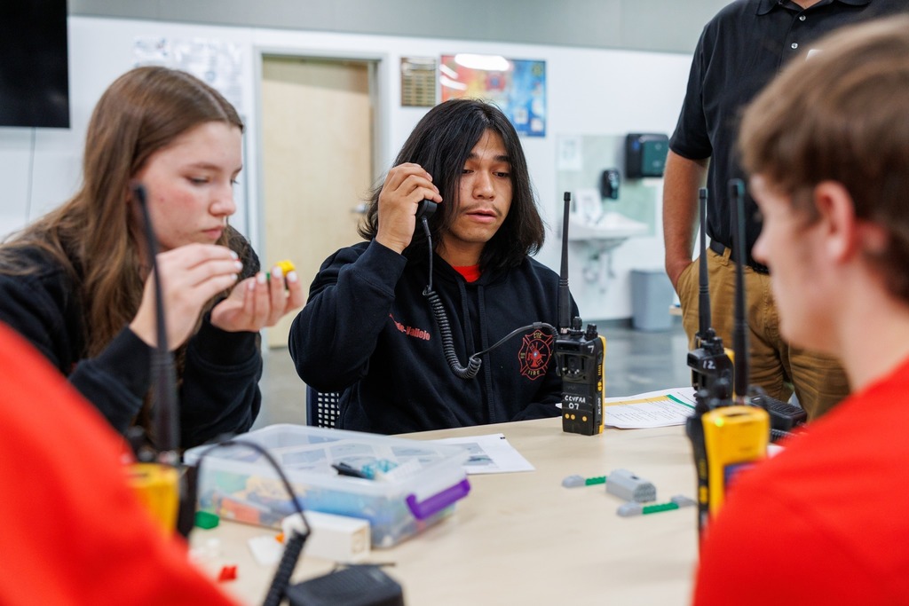 Students sit around a table working with radios and small equipment pieces. One student in a black hoodie with a fire department emblem holds a radio microphone, practicing communication. Another student examines a small object in hand, while others listen and observe. Instruction sheets and supplies are spread across the table.