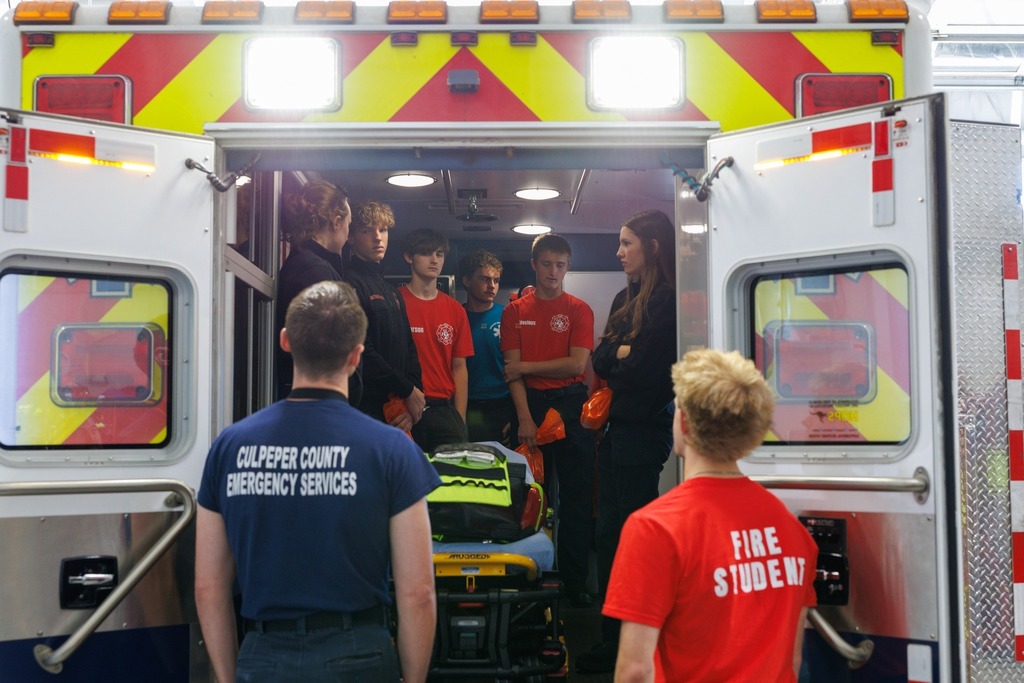 Students in red and blue uniforms stand inside the back of an ambulance, listening to an instructor explain emergency procedures. Two students wearing shirts labeled 'Culpeper County Emergency Services' and 'Fire Student' stand just outside the open ambulance doors. A stretcher is positioned in the center of the ambulance.