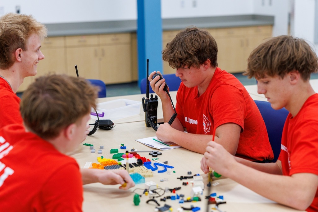 Four students in red fire training shirts sit around a table working on a hands-on activity. One student speaks into a handheld radio while others build with colorful LEGO pieces and small tools. Radios and instruction sheets are also on the table.