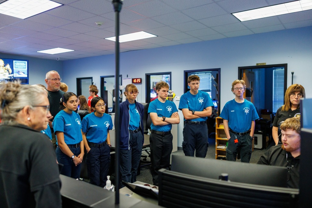 A group of students wearing blue EMS uniforms stand in a dispatch center, listening attentively to an instructor seated at a computer. Two adults stand alongside the students, observing the demonstration. Monitors, office furniture, and wall clocks are visible in the background.