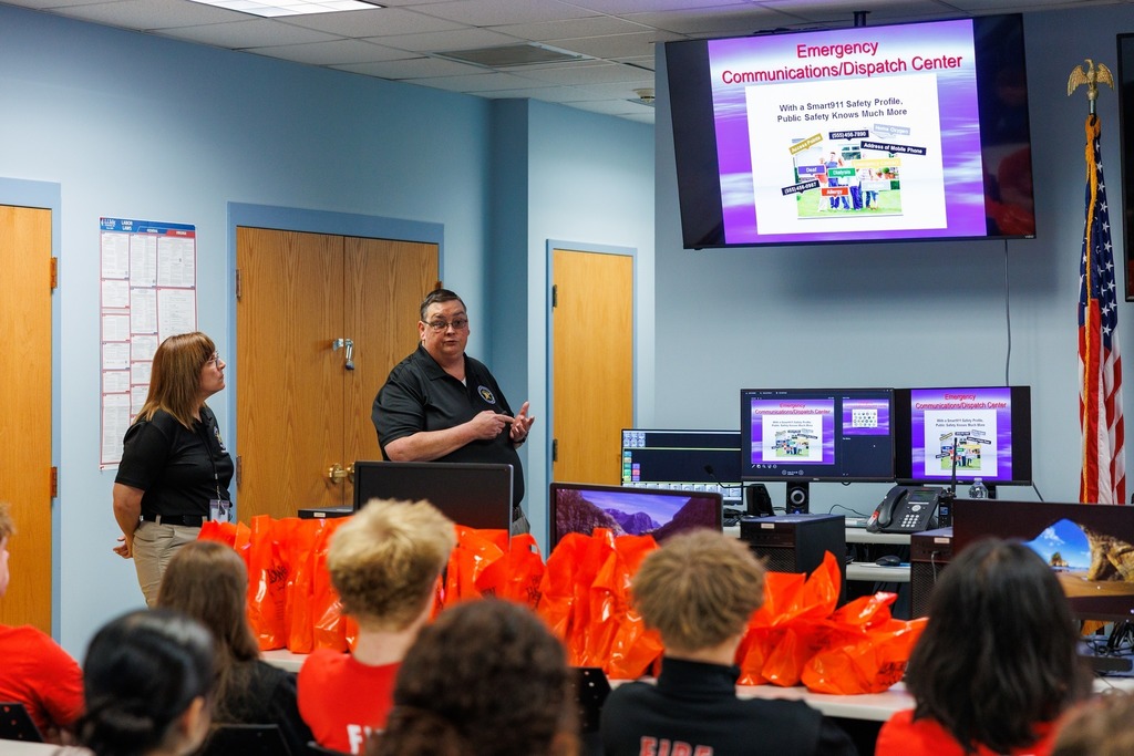 Two presenters in black shirts stand at the front of a classroom, giving a presentation about the Emergency Communications/Dispatch Center. A large screen displays a slide titled 'Emergency Communications/Dispatch Center' with graphics about Smart911 safety profiles. Students in red shirts sit facing forward, with rows of orange tote bags on the desks. Computer monitors and an American flag are also visible in the room.