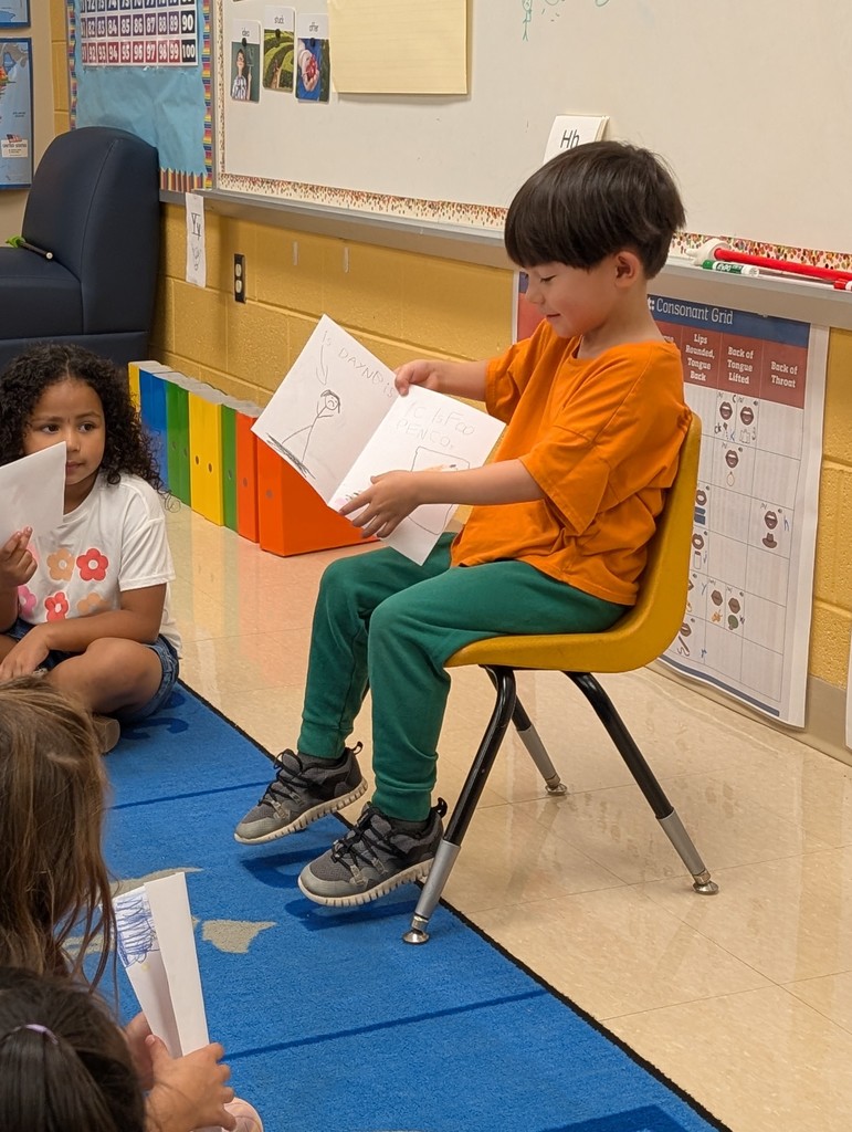 A boy in an orange shirt and green pants sits on a yellow chair at the front of the classroom, holding open his paper with drawings and words. The children on the rug in front of him hold their own papers and look on quietly.